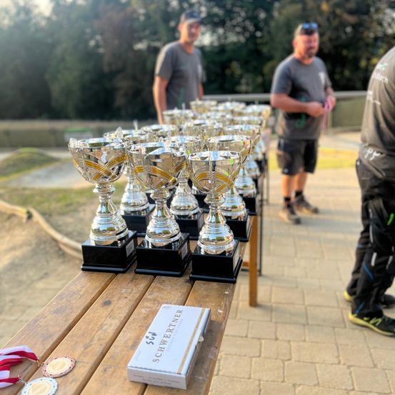 Ein Tisch mit mehreren Silbertrophäen auf einer Ziegelfläche. Ein Kasten mit dem Text Schweitzerner steht auf dem Tisch. Einige Leute stehen hinter dem Tisch.