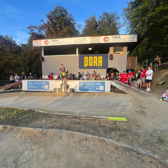 Ein Dirtbike-Rennen mit mehreren Teilnehmern, Bannern und Zuschauern unter einem klaren blauen Himmel. Ein Fahrer steht auf einem Podium mit drei Stufen.