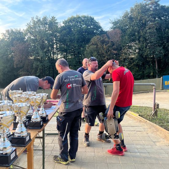 Mehrere Männer stehen um einen Tisch mit Trophäen herum. Ein Mann lässt sich den Kopf von einem anderen rasieren. Ein Mann in einem roten T-Shirt hält ein Skateboard. Bäume und ein Metallzaun sind im Hintergrund zu sehen.