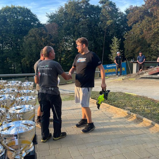 Zwei Männer schütteln sich die Hände vor vielen Trophäen, während andere in der Nähe zusehen. Bäume und ein Banner sind im Hintergrund.