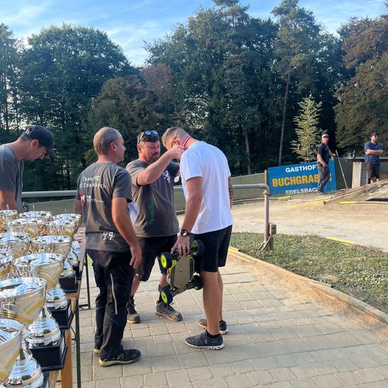 Eine Gruppe von Männern steht bei einem Tisch mit mehreren Trophäen. Ein Mann trägt eine Medaille. Ein Schild steht Gasthof Buchgrab. Bäume und ein Zaun sind im Hintergrund.