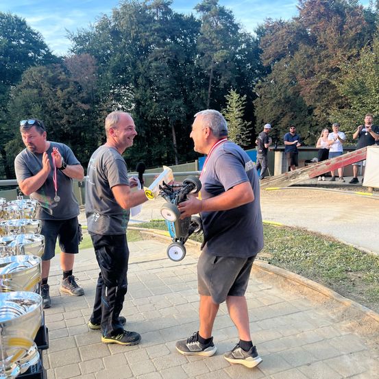 Drei Männer stehen auf einem Feld aus Erde, zwei von ihnen unterhalten sich, während der Dritte einen Pokal hält. Sie tragen alle graue Shirts und Hosen. Hinter ihnen stehen Menschen und beobachten.