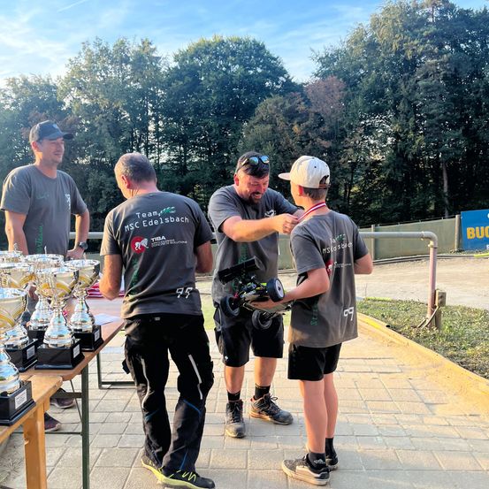 Vier Männer stehen um einen Tisch mit Trophäen. Einer hält ein ferngesteuertes Auto. Dahinter ist ein Banner zu sehen. Bäume und ein bewölkter Himmel bilden den Hintergrund.