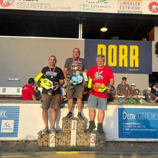 Drei Männer stehen auf einem Podium mit Medaillen und Pokalen. Sie tragen Sonnenbrillen und Freizeitkleidung. Dahinter steht ein Banner mit dem Wort 'DORR' und mehrere Menschen beobachten.