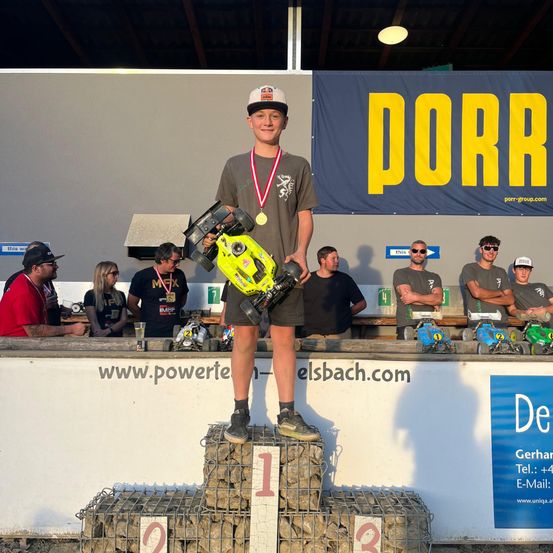 Ein junger Mann steht auf einem Podium und hält ein ferngesteuertes Auto. Er hat eine Medaille um den Hals und trägt eine weiße Kappe. Hinter ihm befindet sich ein Banner mit dem Wort 'PORR' und einer Menge Menschen, die zusehen. Auf dem Podium sind die Zahlen '1', '2' und '3' sichtbar.