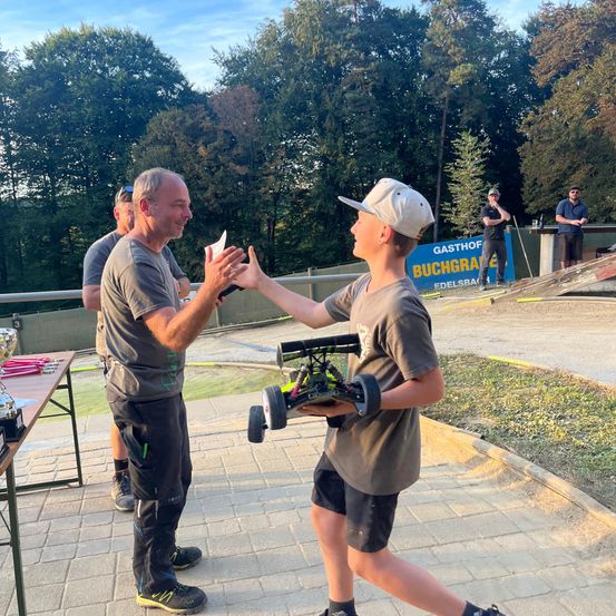 Ein erwachsener Mann überreicht einem jungen Jungen einen Pokal bei einem Dirt-Track-Rennevent. Hinter ihnen steht ein Tisch mit Trophäen und ein Banner ist sichtbar. Mehrere Personen stehen hinter einem Zaun im Hintergrund.