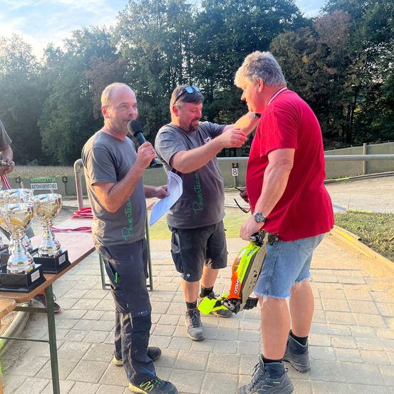 Drei Männer stehen in einem Außenbereich, zwei halten ein Mikrofon und eine Trophäe, der andere eine Kettensäge. Sie befinden sich wahrscheinlich bei einer Veranstaltung. Hinter ihnen steht ein Holztisch mit Trophäen. Bäume und ein Metallzaun sind im Hintergrund.