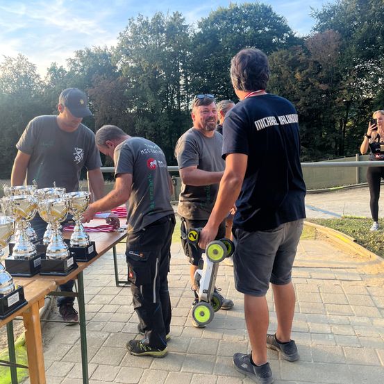 Fünf Männer stehen um einen Tisch mit mehreren Trophäen. Ein Mann hält ein Hoverboard. Hinter ihnen macht eine Person ein Foto. Bäume und ein Zaun sind im Hintergrund.