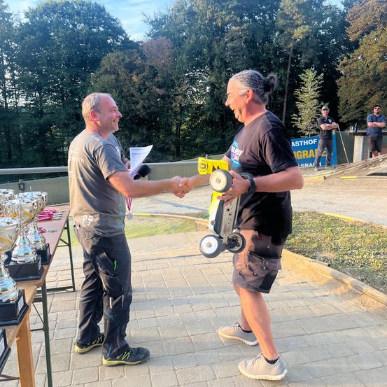 Zwei Männer schütteln sich die Hände bei einer Veranstaltung im Freien. Einer hält eine Trophäe, der andere ein ferngesteuertes Auto. Hinter ihnen steht ein Tisch mit Trophäen und zwei Männer in Freizeitkleidung. Bäume und ein Banner im Hintergrund.