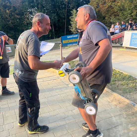 Zwei Männer schütteln sich die Hände und halten ein ferngesteuertes Auto, tragen Medaillen. Ein Mann mit einer Medaille an seinem Shirt hält ein Papier. Im Hintergrund steht eine Gruppe von Menschen auf einer Plattform mit Bannern.