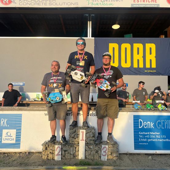 Drei Männer stehen mit Spielzeugautos auf einem Podest und tragen Medaillen. Sie lächeln. Das Podest hat die Zahlen 1, 2 und 3. Dahinter hängt ein Banner mit dem Wort Dorr.