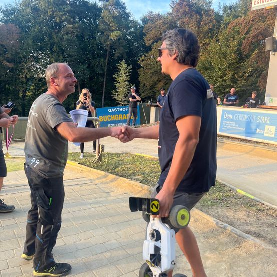 Zwei Männer schütteln sich die Hände vor einer Menschenmenge, einer mit einer Medaille und der andere fährt einen Roller. Hinter ihnen befindet sich ein Banner mit deutschem Text.
