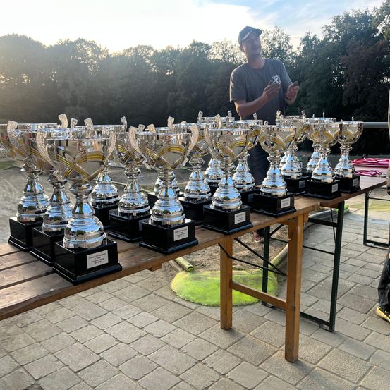 Ein Mann steht hinter einem Tisch voller Trophäen, mit Bäumen im Hintergrund.