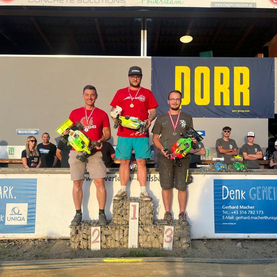 Drei Männer stehen mit Medaillen und Spielzeugautos lächelnd auf einem Podest. Dahinter beobachten andere. Ein Banner sagt 'DORR' und 'Denk GEMEIN'. Die Umgebung scheint ein Wettbewerbsort zu sein.