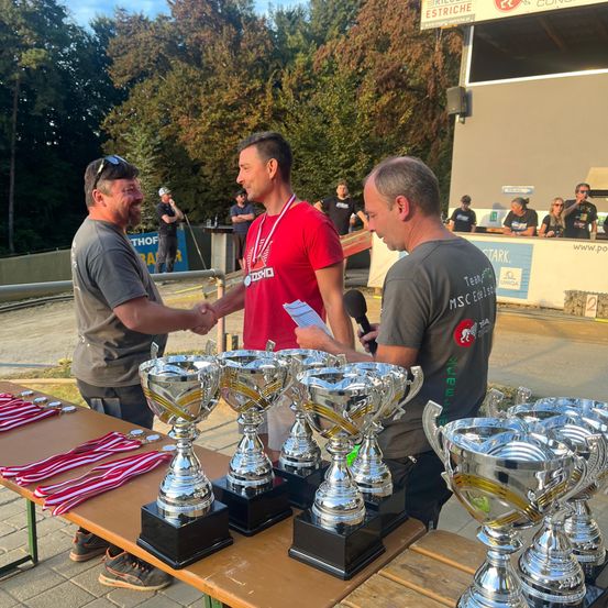Drei Männer stehen hinter einem Tisch mit Trophäen. Einer hält ein Mikrofon und ein anderer schüttelt einem Mann die Hand. Dahinter sitzen mehrere Personen auf Bänken und beobachten. Bäume und ein Gebäude sind im Hintergrund.