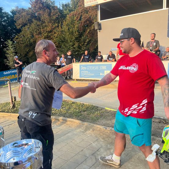 Zwei Männer schütteln sich im Freien bei einer Veranstaltung die Hände, mit einem Banner hinter ihnen. Ein Mann trägt ein rotes Shirt und blaue Shorts, der andere ein graues Shirt mit einer Nummer darauf.