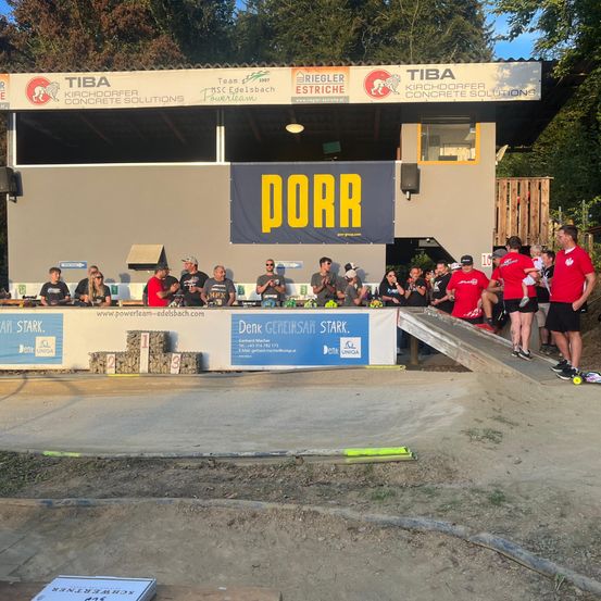 Eine Gruppe von Menschen steht vor einer Bühne mit einem Banner, auf dem 'PORR' steht. Sie tragen rote Shirts und schwarze Shorts. Die Bühne hat einen Lautsprecher und eine Lampe.