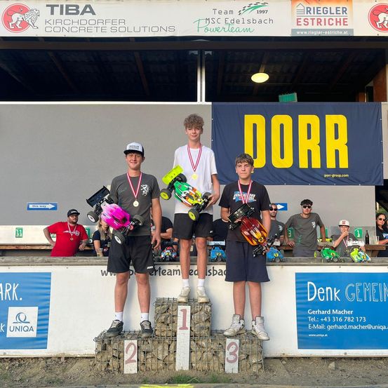 Drei Rennfahrer stehen auf einem Podium mit den Nummern 1, 2 und 3 und halten Trophäen. Sie tragen Medaillen und Rennanzüge. Hinter ihnen steht auf einem Banner 'DORR'. Mehrere Leute schauen im Hintergrund zu, einige tragen Sonnenbrillen und Hüte. Werbebanner und ein Logo sind an der Wand.