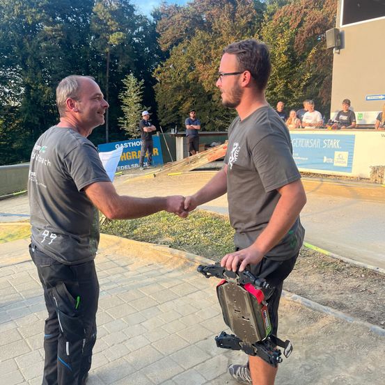 Zwei Männer schütteln sich die Hände auf einem gepflasterten Weg. Dahinter hält eine Person ein Skateboard mit einem roten und schwarzen Design. Im Hintergrund steht eine Gruppe von Menschen in der Nähe eines Banners mit Text und einem Lautsprecher. Bäume und ein Gebäude sind sichtbar.