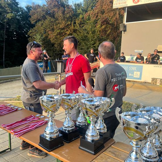 Ein Mann in einem roten T-Shirt schüttelt die Hand eines anderen Mannes, während ein dritter Mann hinter ihnen steht, alle vor einem Tisch mit mehreren Pokalen. Im Hintergrund befinden sich ein Zaun, Bäume und ein Gebäude.