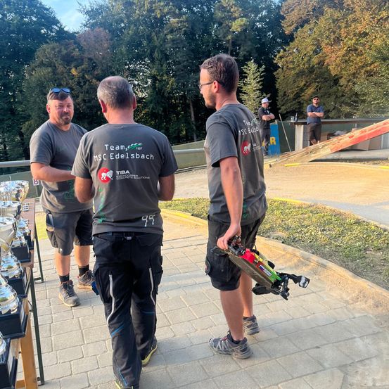 Drei Männer gehen in einem Park mit einer Rampe und Trophäen im Hintergrund. Sie tragen graue T-Shirts mit dem Team MSC Edelsbach Logo. Ein Mann hält ein Spielzeugauto.