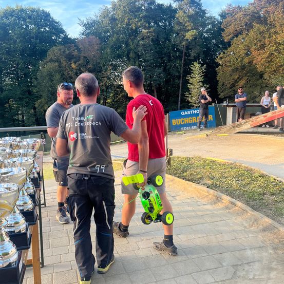 Drei Männer gehen an einer Trophäen-Ausstellung vorbei, einer hält ein Spielzeugauto. Dahinter beobachten Leute von einer Rampe.
