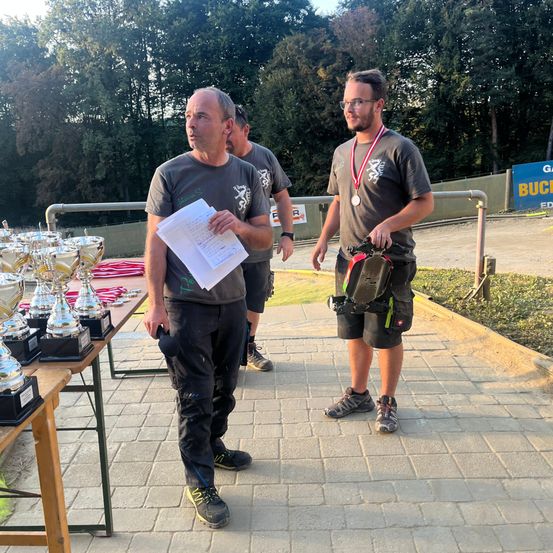Zwei Männer stehen auf einem gepflasterten Weg mit Trophäen auf einem Tisch. Einer hält ein Papier und der andere trägt eine Medaille. Hinter ihnen ist ein verschwommener Hintergrund mit Bäumen und einem blauen Banner.