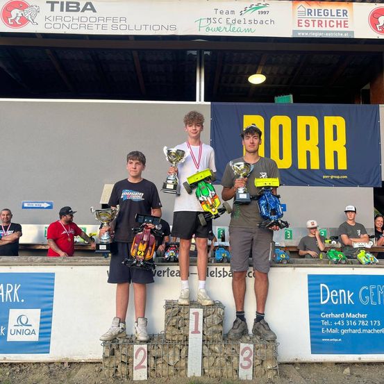 Drei junge Männer stehen mit Pokalen in der Hand auf einem Podest. Hinter ihnen steht ein Banner mit der Aufschrift 'DORR'. Zuschauer und Personen im Hintergrund tragen Mützen und Turnschuhe.