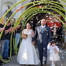 Ein Hochzeitspaar und ein junges Mädchen gehen durch einen grünen Bogen mit fallenden weißen Blütenblättern. Der Bräutigam trägt einen blauen Anzug und die Braut ein weißes Kleid. Die Leute halten Blumen.