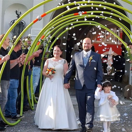 Ein Hochzeitspaar und ein junges Mädchen gehen durch einen grünen Bogen mit fallenden weißen Blütenblättern. Der Bräutigam trägt einen blauen Anzug und die Braut ein weißes Kleid. Die Leute halten Blumen.