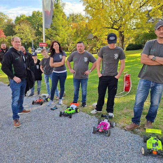 Eine Gruppe von Menschen steht auf einem Schotterweg mit ferngesteuerten Autos. Sie sind lässig gekleidet und lächeln. Hinter ihnen befindet sich ein Rasenbereich mit Bäumen und einem Feuerlöscher.