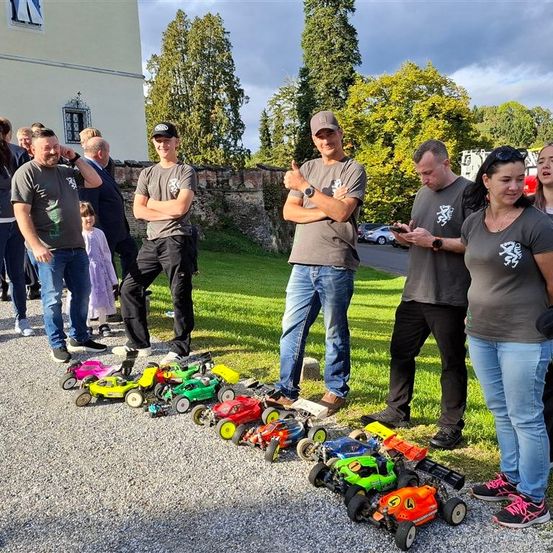 Eine Gruppe von Menschen steht auf einem Kiesweg mit ausgestellten Spielzeugautos. Eine Frau schaut auf ihr Handy. Dahinter steht ein Gebäude mit Bäumen und geparkten Autos.