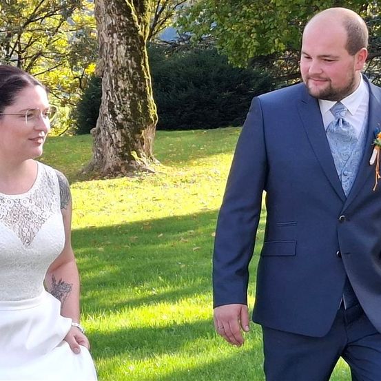 Ein Mann und eine Frau in formeller Kleidung stehen auf einem Rasen und posieren lächelnd für ein Foto. Der Mann trägt Anzug und Krawatte mit einer Blume, die Frau ein weißes Kleid und eine Brille. Dahinter steht ein Baumstamm und einige Büsche.