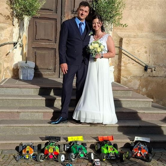 Ein frisch verheiratetes Paar steht auf Stufen mit mehreren Spielzeugautos, einem Gebäude im Hintergrund und Topfpflanzen.