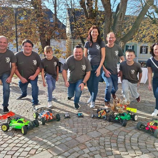 Eine Gruppe von Menschen in passenden T-Shirts steht vor einem Baum und lächelt für ein Foto. Verschiedene Spielzeugautos sind auf dem gepflasterten Boden vor ihnen platziert.