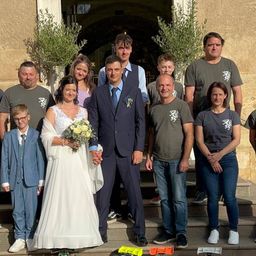Eine Gruppe von Menschen in formeller Kleidung steht auf den Stufen eines Gebäudes, wahrscheinlich für eine Hochzeit. Die Braut und der Bräutigam stehen in der Mitte, umgeben von Familienmitgliedern.