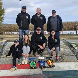 Eine Gruppe von Menschen posiert für ein Foto vor einem Maisfeld mit zwei Modellen von Rennwagen vor ihnen.