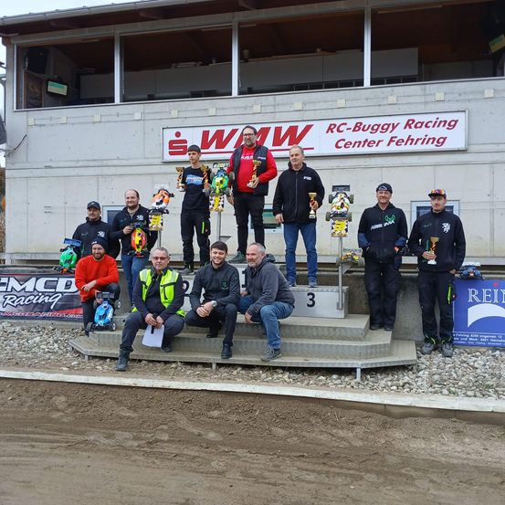 Eine Gruppe von Menschen steht mit Trophäen auf einem Podium und trägt Winterkleidung. Ein Banner mit der Aufschrift RC-Buggy Racing Center Fehring ist an der Wand montiert. Sie stehen auf einer Betonfläche mit einigen Steinen.