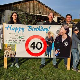 Eine Gruppe von Menschen steht vor einem Haus, lächelt und posiert für ein Foto. Sie halten ein Schild, auf dem 'Happy Birthday!' steht und ein Geschwindigkeitsbegrenzungsschild für 40. Einige tragen Freizeitkleidung.