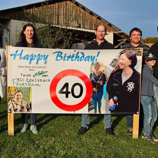 Eine Gruppe von Menschen steht vor einem Haus, lächelt und posiert für ein Foto. Sie halten ein Schild, auf dem 'Happy Birthday!' steht und ein Geschwindigkeitsbegrenzungsschild für 40. Einige tragen Freizeitkleidung.
