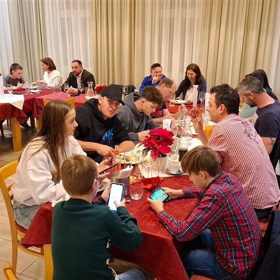 Eine Gruppe von Menschen sitzt an einem Esstisch, einige benutzen ihre Handys. Der Tisch ist mit Tellern, Gläsern und Blumen gedeckt. Im Hintergrund befinden sich Vorhänge.