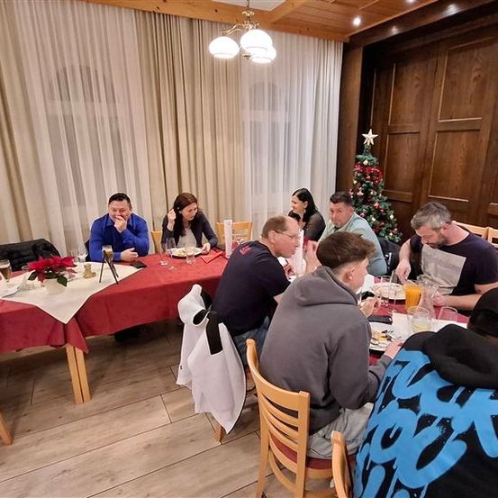 Eine Gruppe von Menschen sitzt um einen Tisch in einem Esszimmer und genießt ihre Mahlzeit. Der Tisch ist mit einer roten Tischdecke bedeckt, und der Raum hat Holzwände und -böden. Eine Weihnachtsbaum steht in der Ecke.