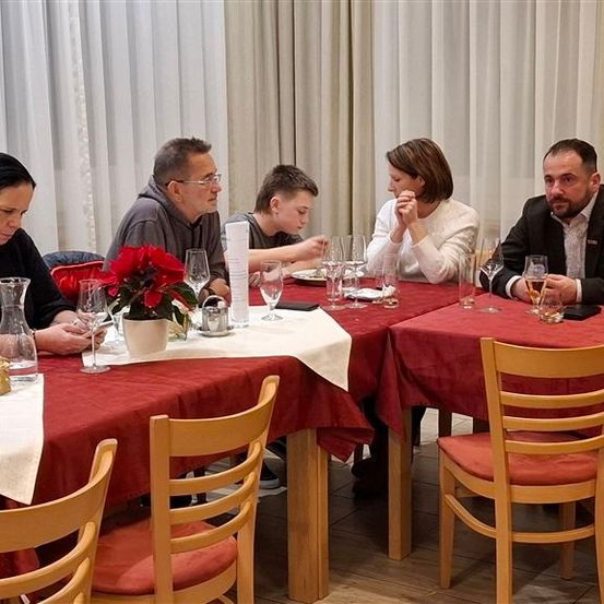 Eine Familie sitzt an einem rot belegten Tisch in einem Restaurant, wobei jedes Familienmitglied in einem Moment der Stille vertieft ist. Gläser, Teller und eine Vase mit roten Blumen stehen auf dem Tisch. Der Raum ist mit weißen Vorhängen dekoriert.