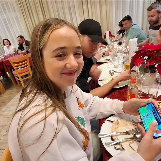 Ein Familienessen mit einer Frau in einem festlichen Shirt, die ein Selfie mit ihrem Handy macht. Sie sitzt an einem Tisch mit einem Teller Essen. Hinter ihr sitzen Menschen, essen und unterhalten sich. Im Vordergrund steht eine rote Weihnachtsblume.