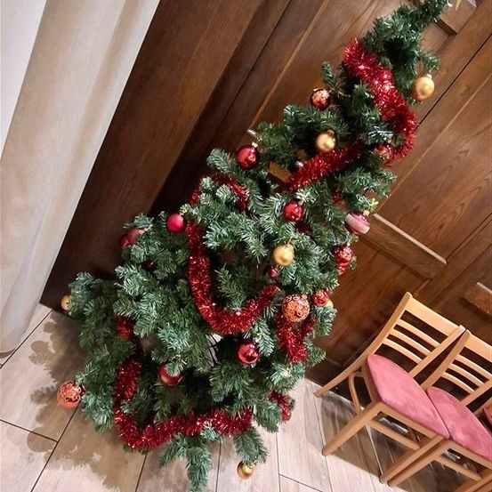 Ein mit roten und goldenen Ornamenten geschmückter Weihnachtsbaum, verziert mit einem Stern, steht geneigt an einer Holztür. In der Nähe befinden sich zwei Holzstühle mit rosa Kissen auf einem gefliesten Boden.