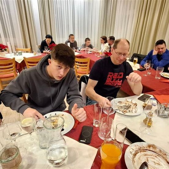 Eine Gruppe von Menschen sitzt an einem Tisch in einem Restaurant. Einige von ihnen essen, während andere auf ihre Handys schauen. Der Tisch ist mit Tellern, Gläsern und einem Handy gedeckt. Die Person in der Mitte trägt ein schwarzes Shirt mit einem Red Bull-Logo.