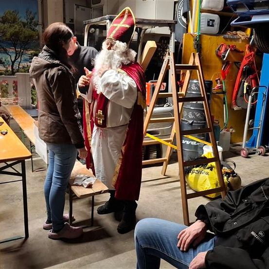 Ein Mann in einem Weihnachtsmannkostüm steht neben einer Bank mit einer anderen Person, während andere zusehen. In der Nähe wird an einem Auto gearbeitet.