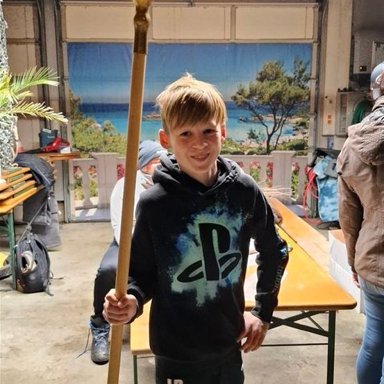 Ein Junge in einer Kapuzenjacke steht in einer Garage, hält einen Stab und um ihn herum Leute und Möbel. Hinter ihm ein Tisch mit einem Rucksack.