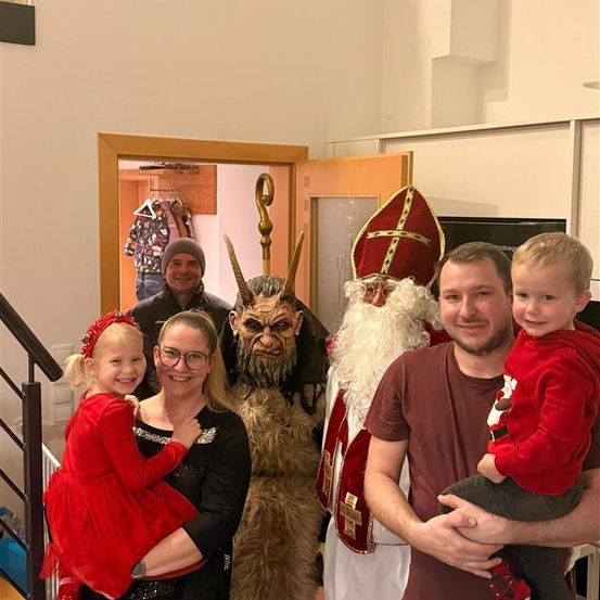 Eine Familie posiert für ein Foto mit einer Person als St. Nikolaus verkleidet und einem Teufel im Wohnzimmer.