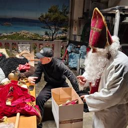 Drei Männer sind in einem Raum. Einer trägt einen Weihnachtsmann-Kostüm. Ein anderer ist in einer schwarzen Jacke und Kappe. Der dritte ist in einem grauen Sweatshirt. Sie sitzen um einen Tisch mit Kostümen und Requisiten.